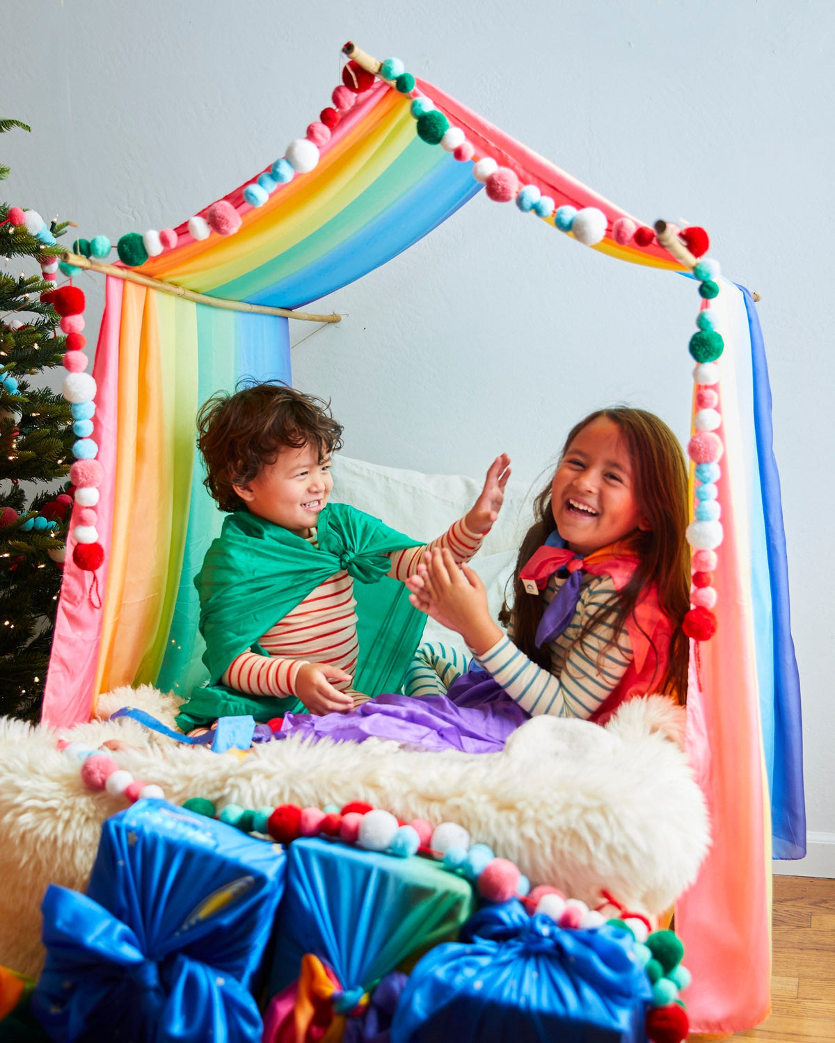 Two children sitting inside a colorful Christmas canopy with Christmas Tree and gifts seen outside sarasilk