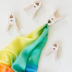 Four wooden clips hold a rainbow silk scarf against a white background, inviting creative play with open-ended toys like Fort Kits.